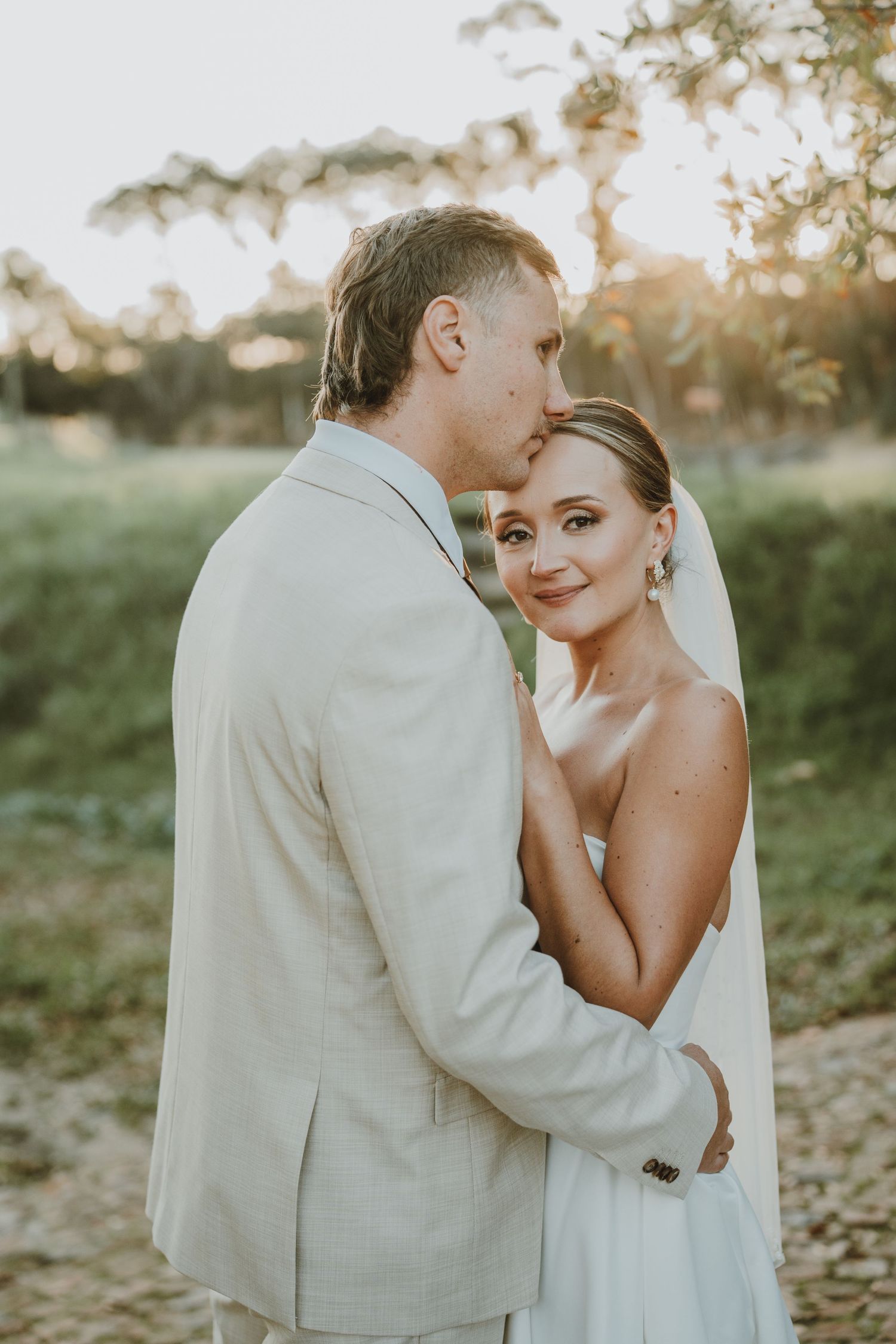 A bride and groom embrace outdoors at golden hour, she wears a white gown and veil, he wears a light beige suit.