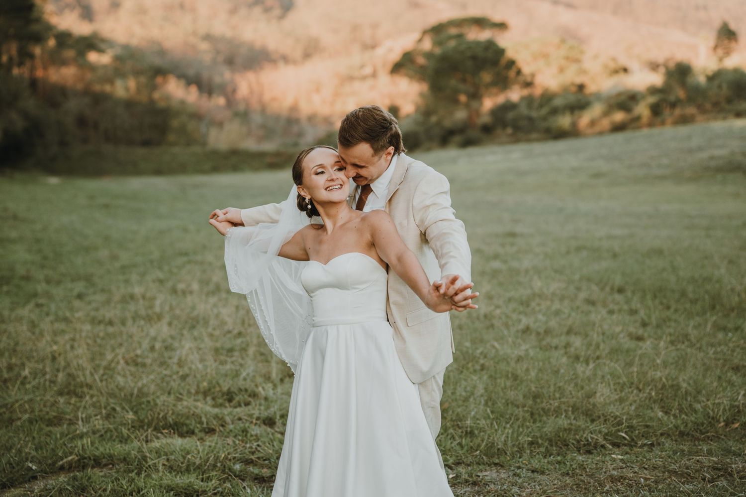 Bride in white gown and groom in tan suit embrace and dance in a lush green field with rolling hills at golden hour.
