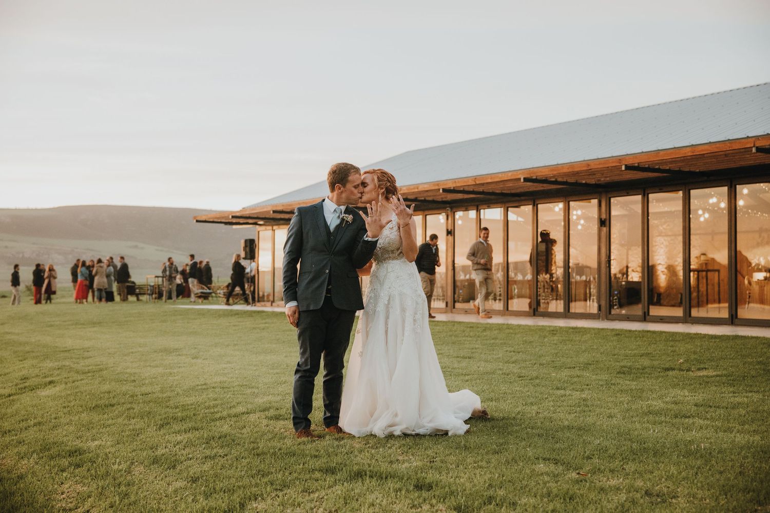 Bride and groom share a kiss on green lawn at sunset outside a modern glass wedding venue.
