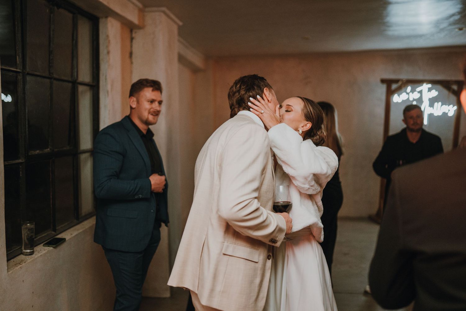 Bride and groom share a first kiss at their wedding reception while guests look on in a warmly lit venue.