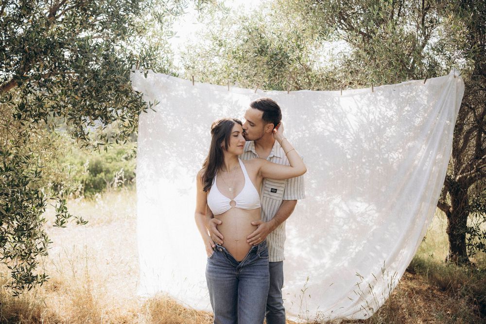 A romantic couple shares tender moments in an olive grove with soft natural lighting and a flowing white fabric backdrop.