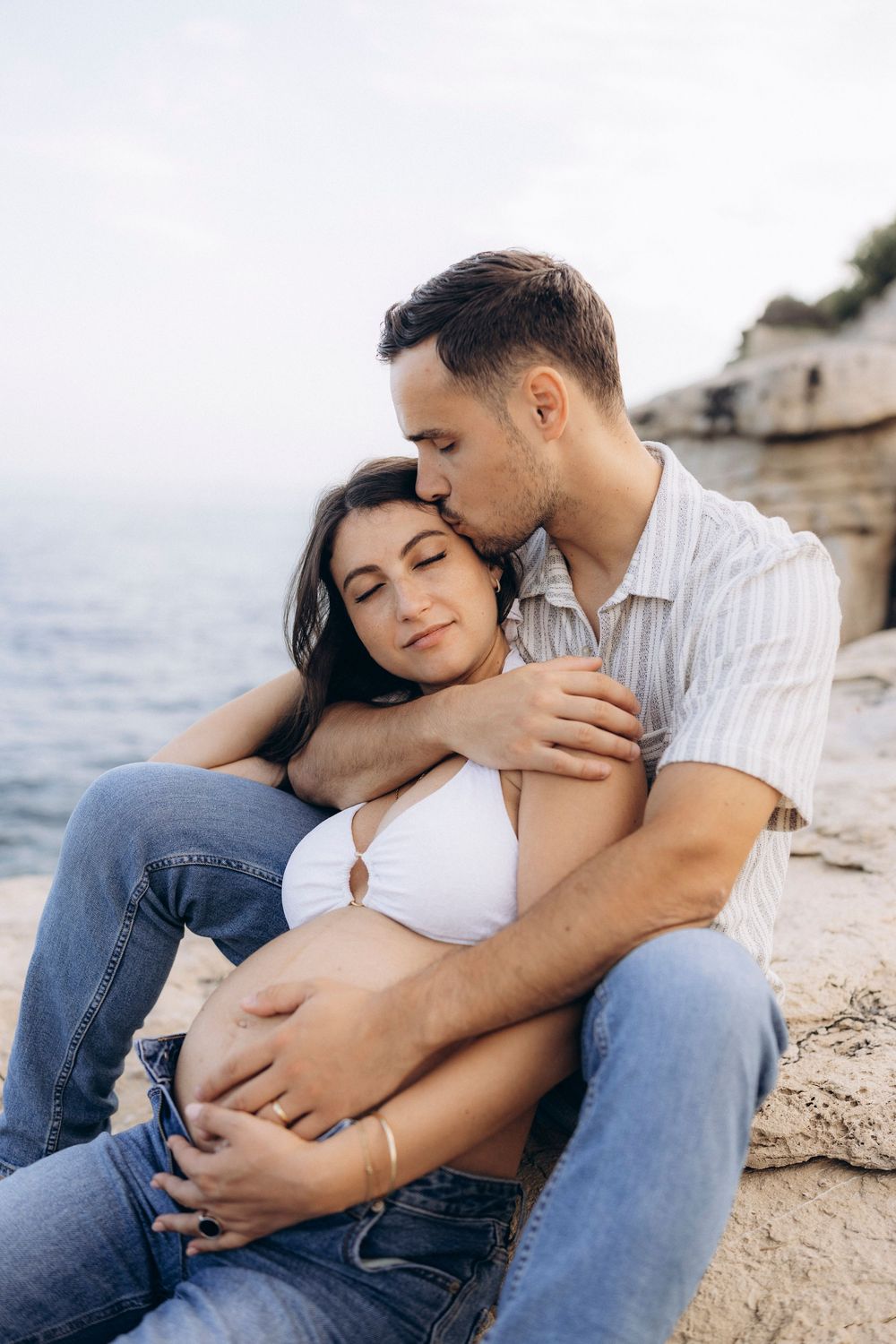 A romantic couple embraces tenderly on a rocky beach during a sunset photoshoot.