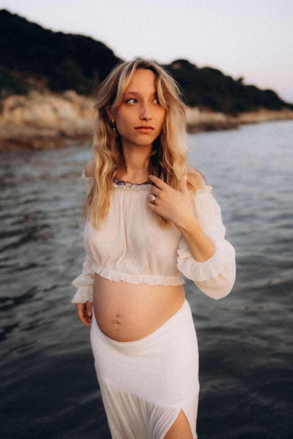 A figure in a white flowing dress poses against rocky coastal waters at sunset creating a dreamy seaside portrait.