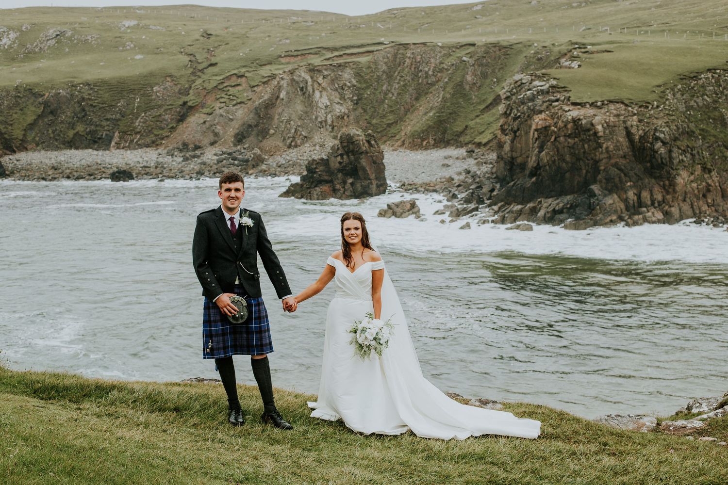 A wedding couple sitting on an old stone wall overlooking Scottish coastal landscape.