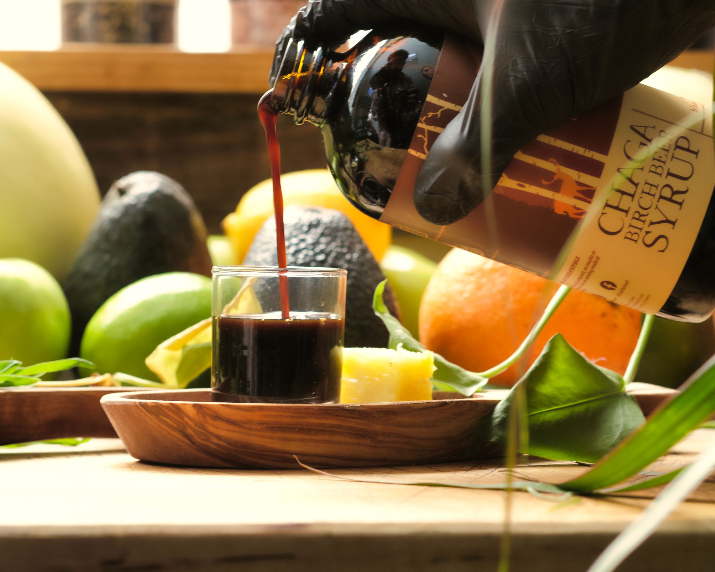 Dark liquid being poured into wooden bowl surrounded by fresh citrus fruits.