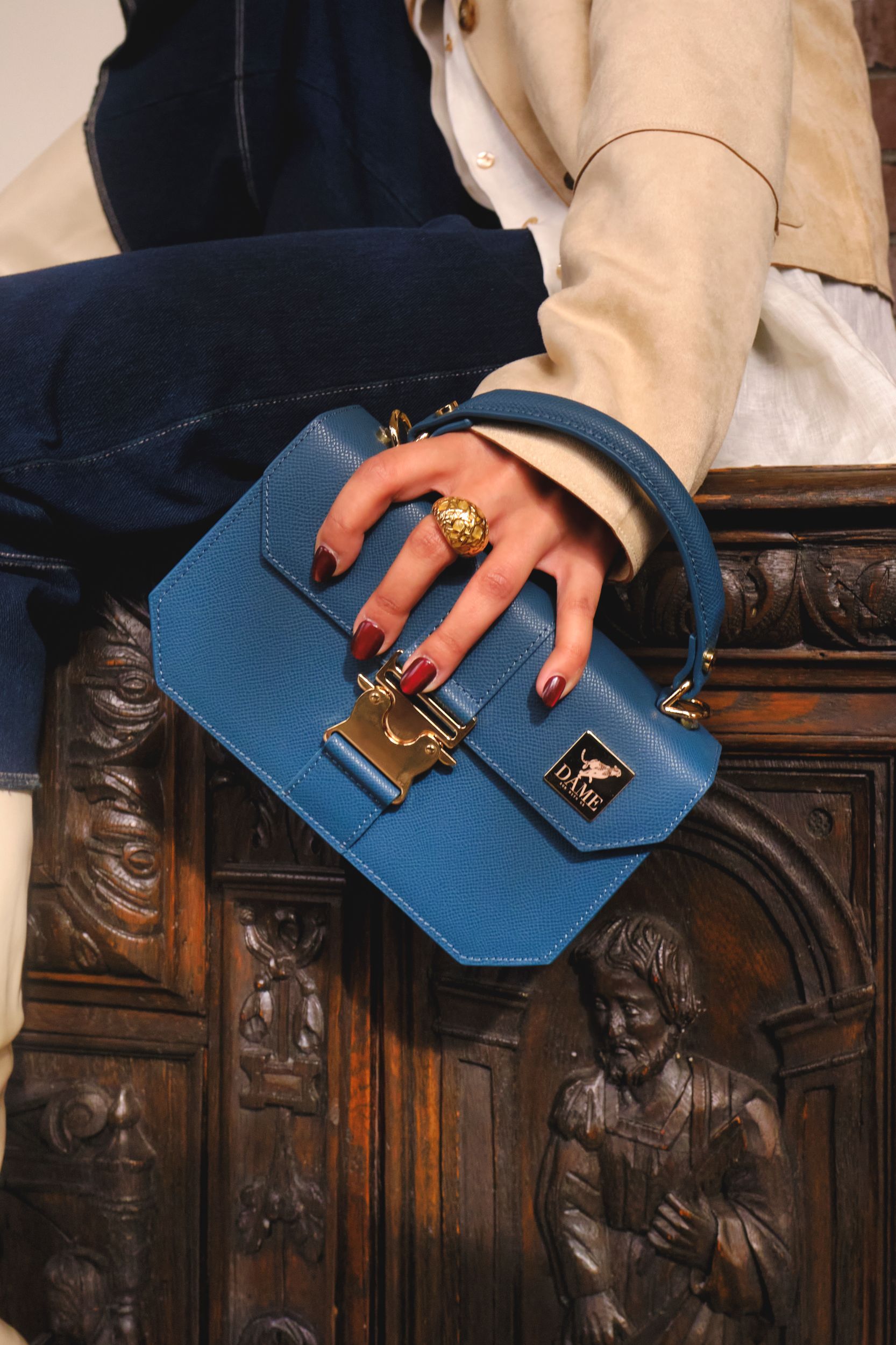 A hand wearing gold rings holds a small blue structured handbag against an ornate wooden carved background.