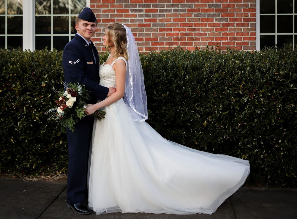Military groom in dress uniform holding bouquet beside bride in white gown and veil outdoors.
