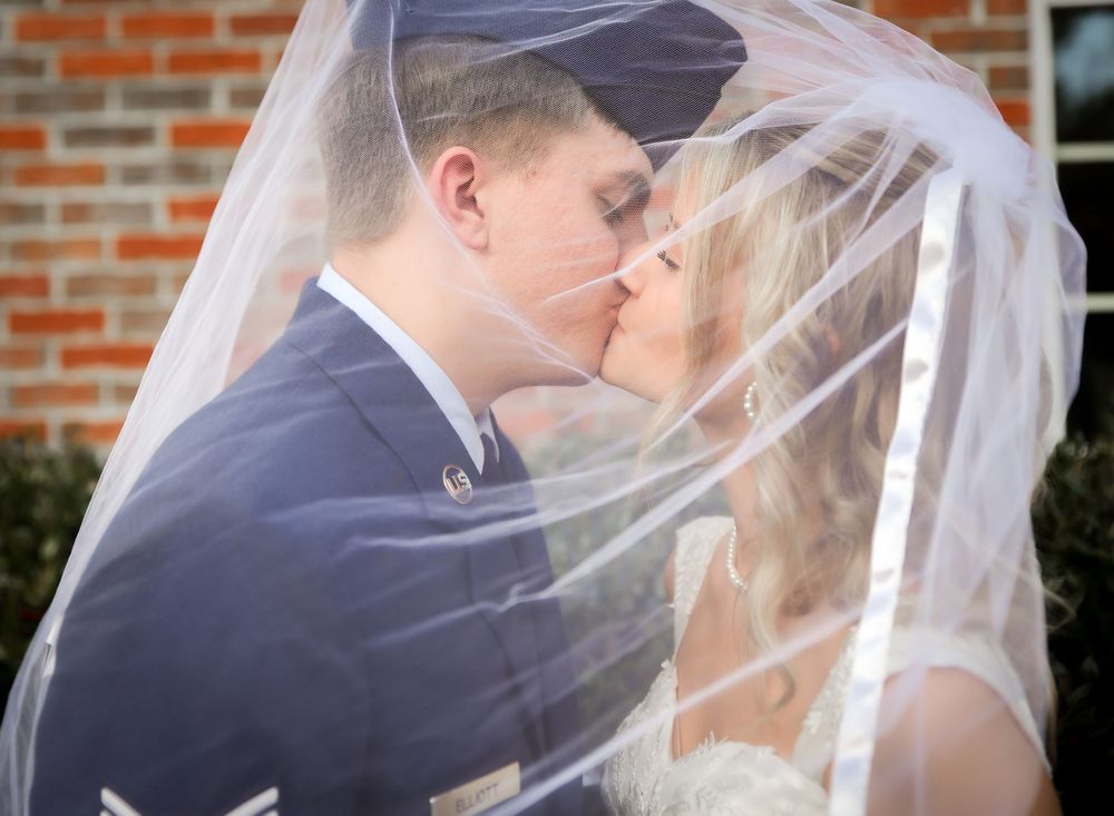 Bride and groom sharing a kiss under flowing white wedding veil near brick wall.