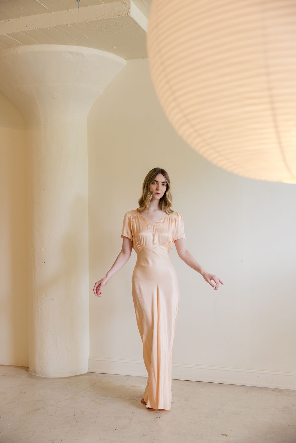 Woman in peach satin maxi dress with short sleeves poses in minimalist white room with column and paper lantern.