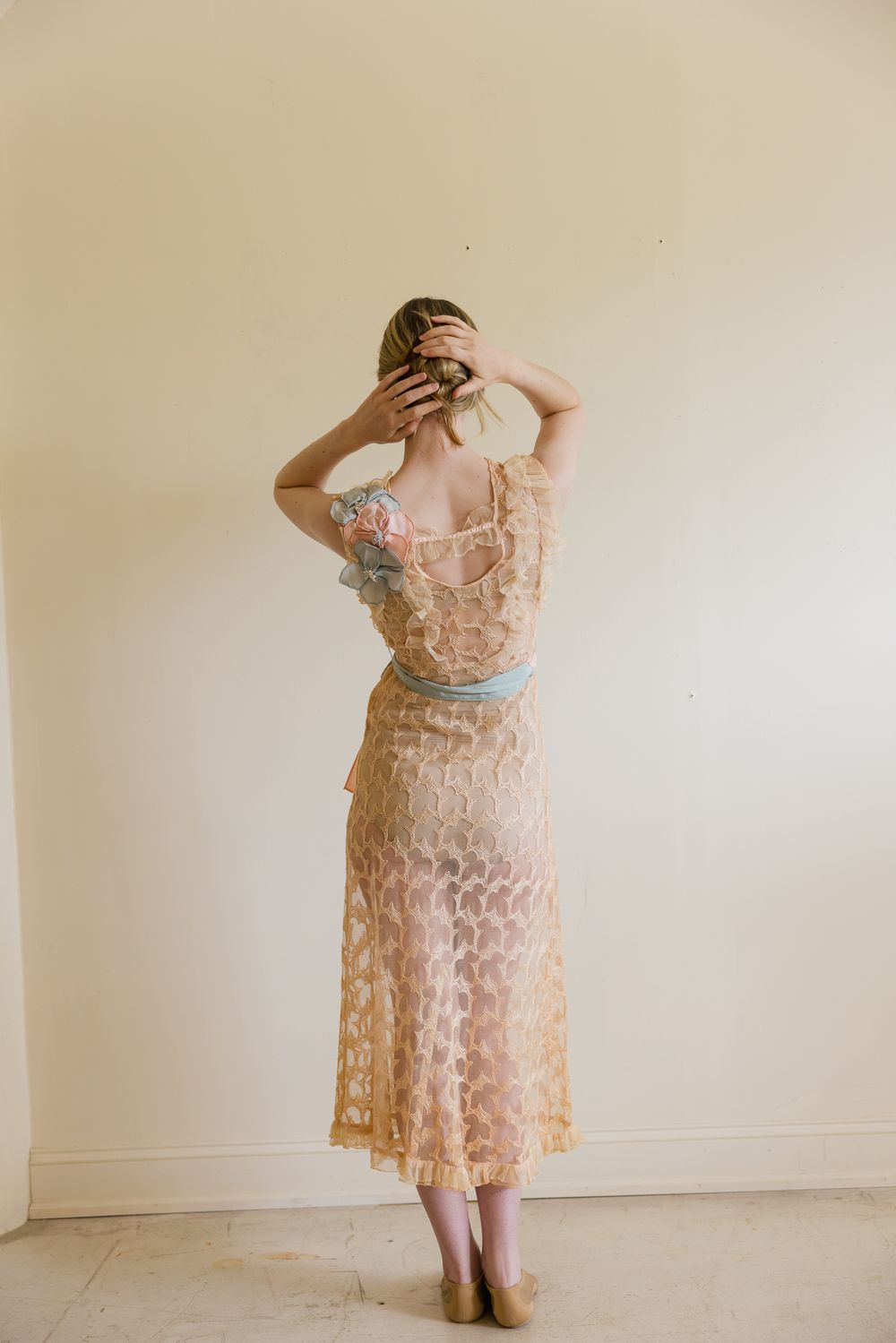 Woman in sheer peach lace midi dress with blue trim, standing with back to camera, hands in hair.