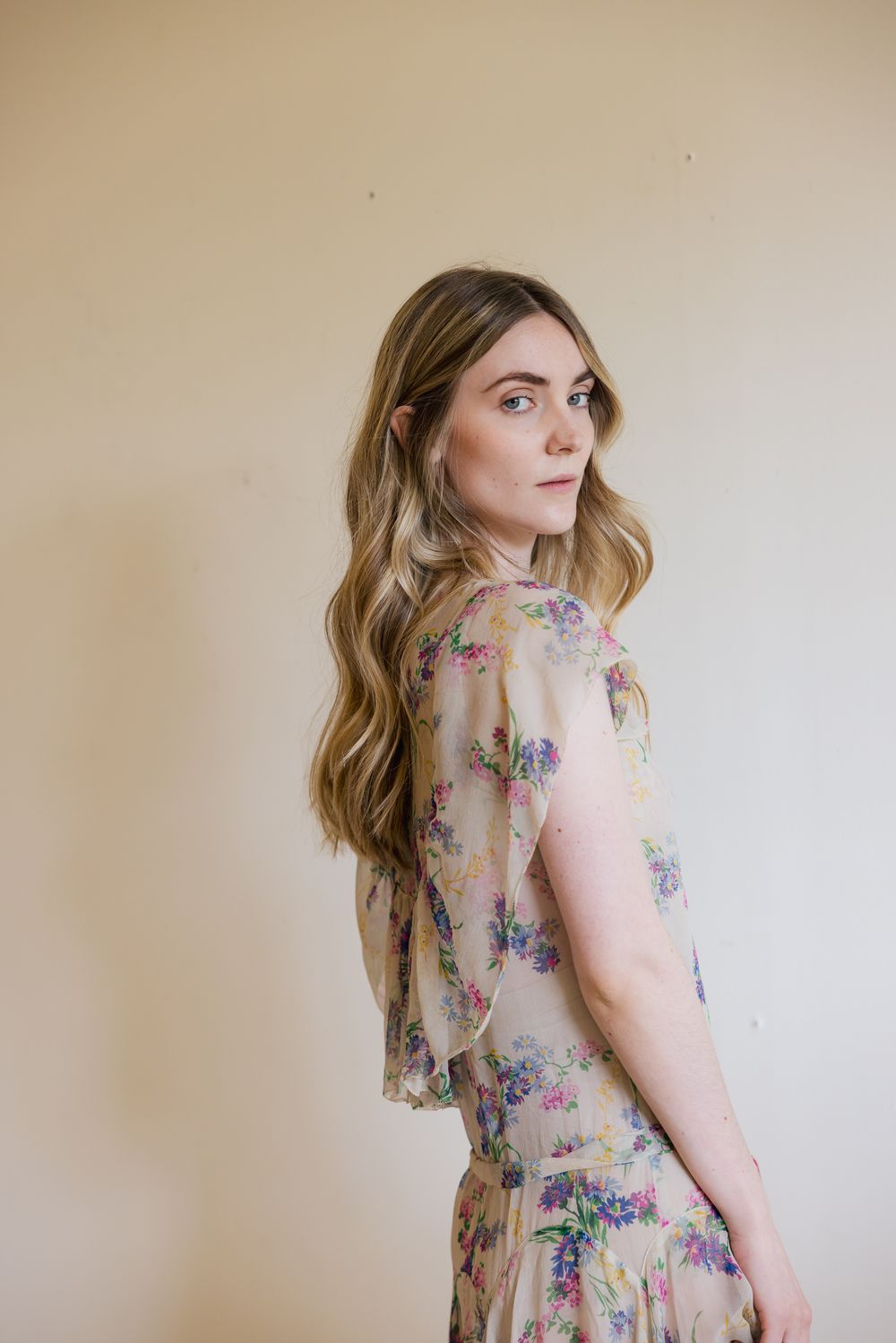 Woman in floral chiffon dress with wavy blonde hair poses sideways, glancing over shoulder against beige wall.