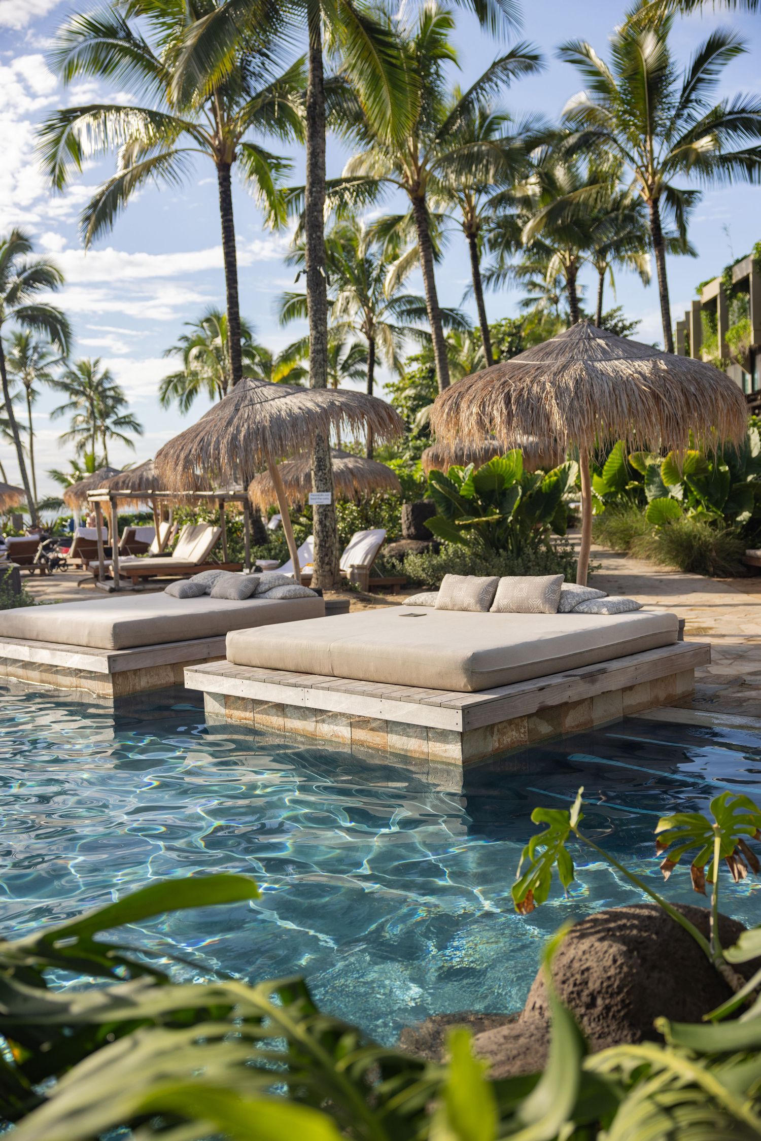 Resort pool with thatched umbrellas and lounge chairs surrounded by palm trees.