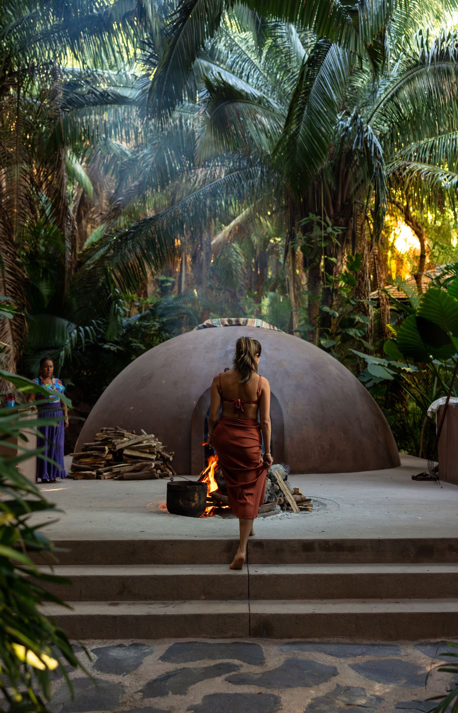 A sweat lodge ceremony setup with dome-shaped structures surrounded by tropical palm trees in a sacred space at sunset.