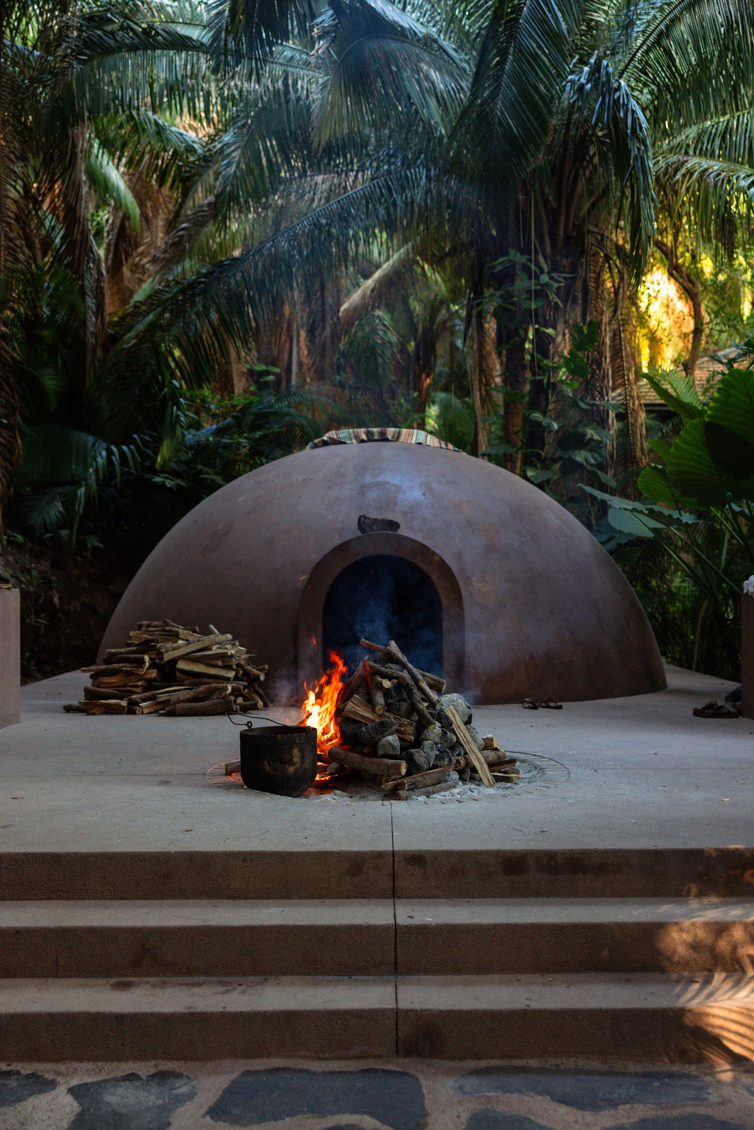 Traditional clay dome structure with burning fire pit against tropical palm tree backdrop at sunset.