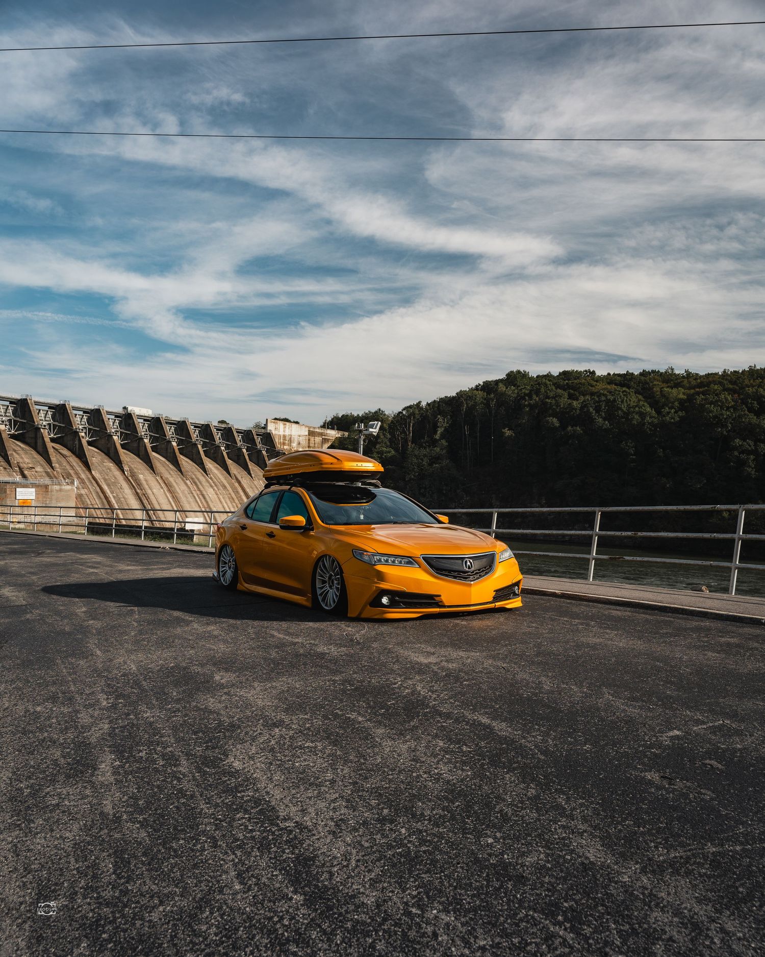 Orange sports car parked near concrete dam structure under dramatic cloudy sky.