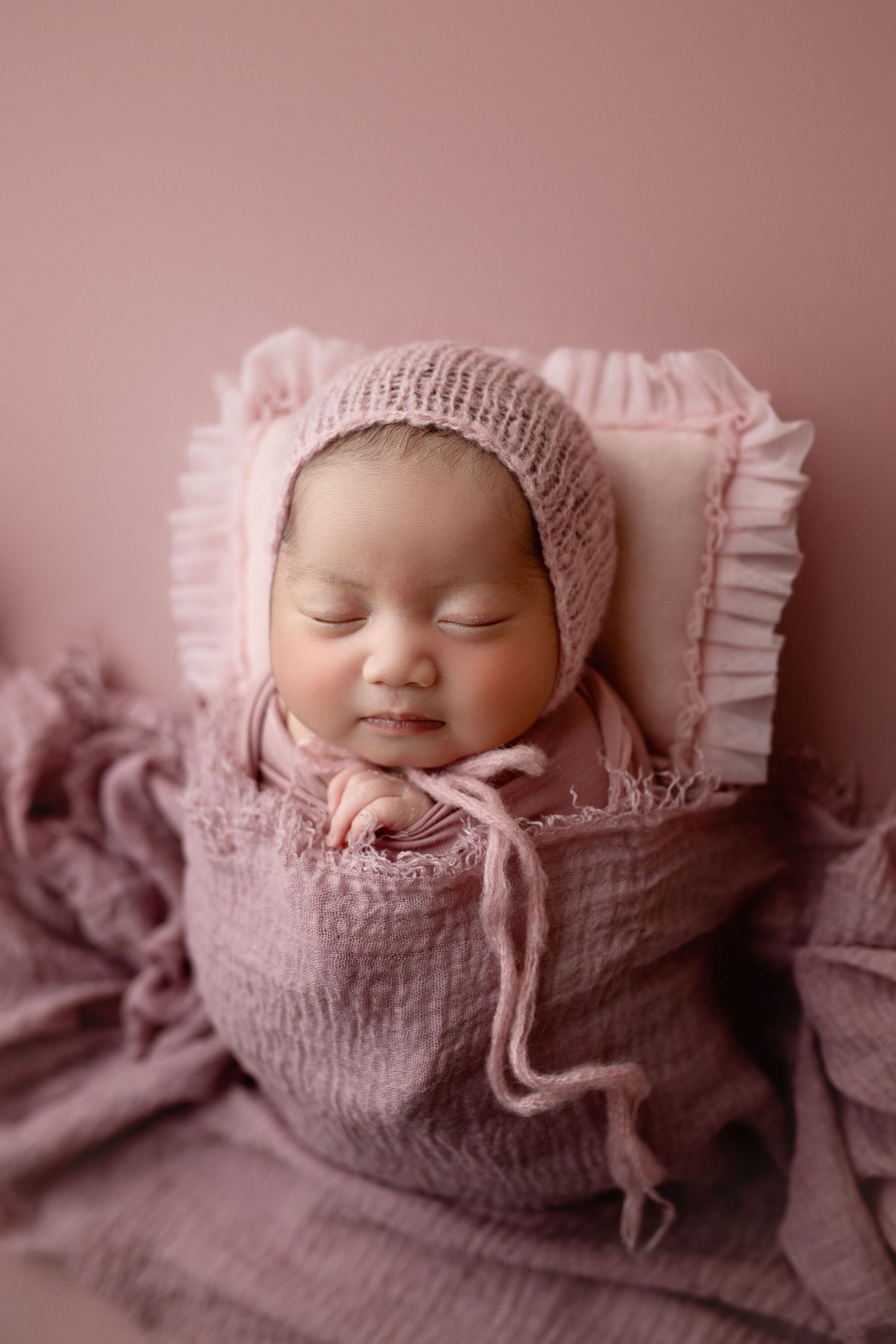 Peaceful sleeping baby in pink knit bonnet and wrap on ruffled pillow.