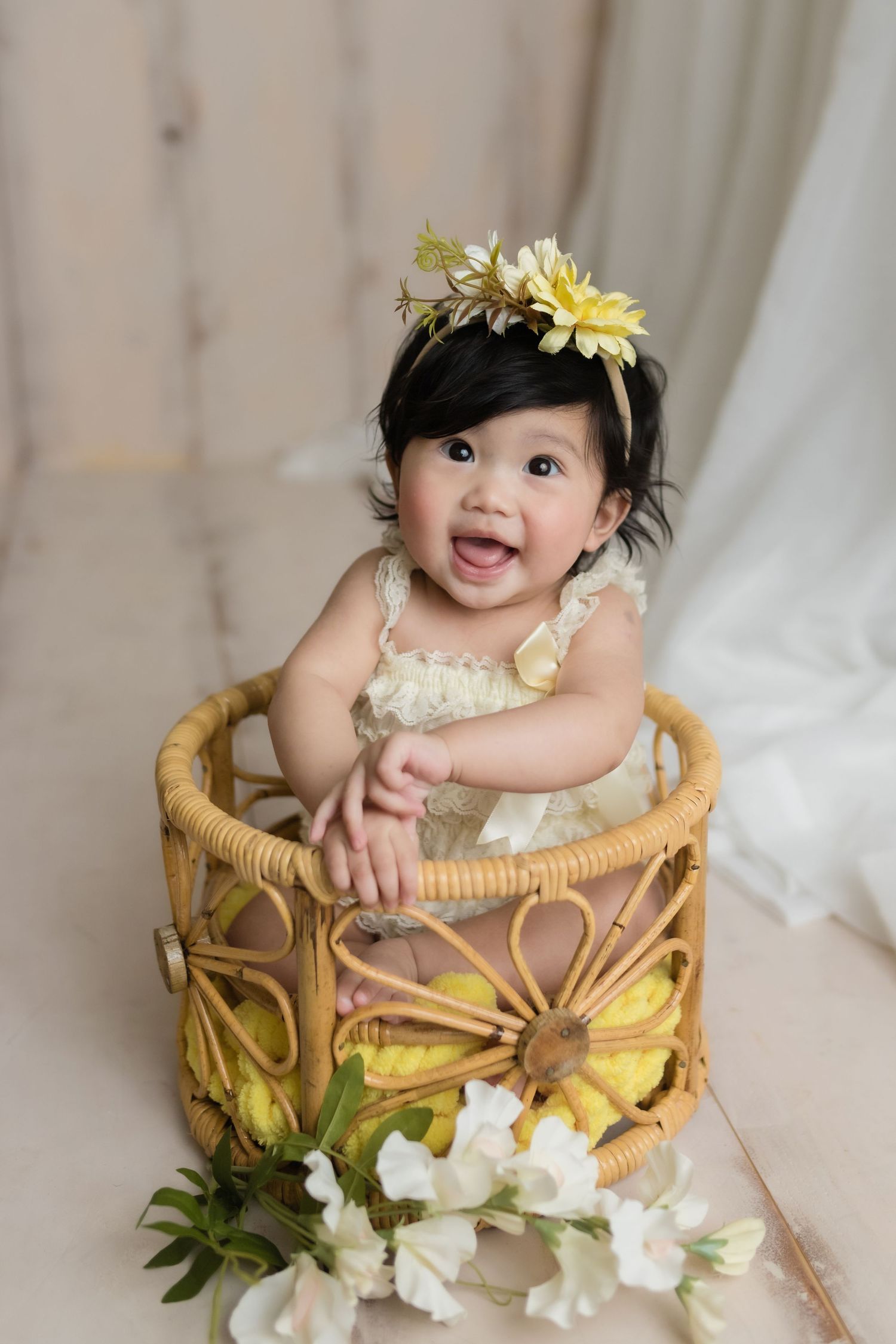 Baby sitting in woven basket wearing flower crown and white outfit against light backdrop.