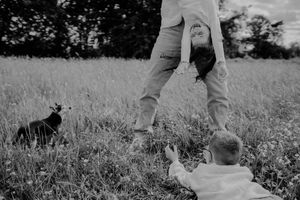 Ein Papa hält seine Tochter über Kopf und die Haare und Arme hängen lang runter. Sie lacht herzhaft. Der Bruder liegt davor im Gras.