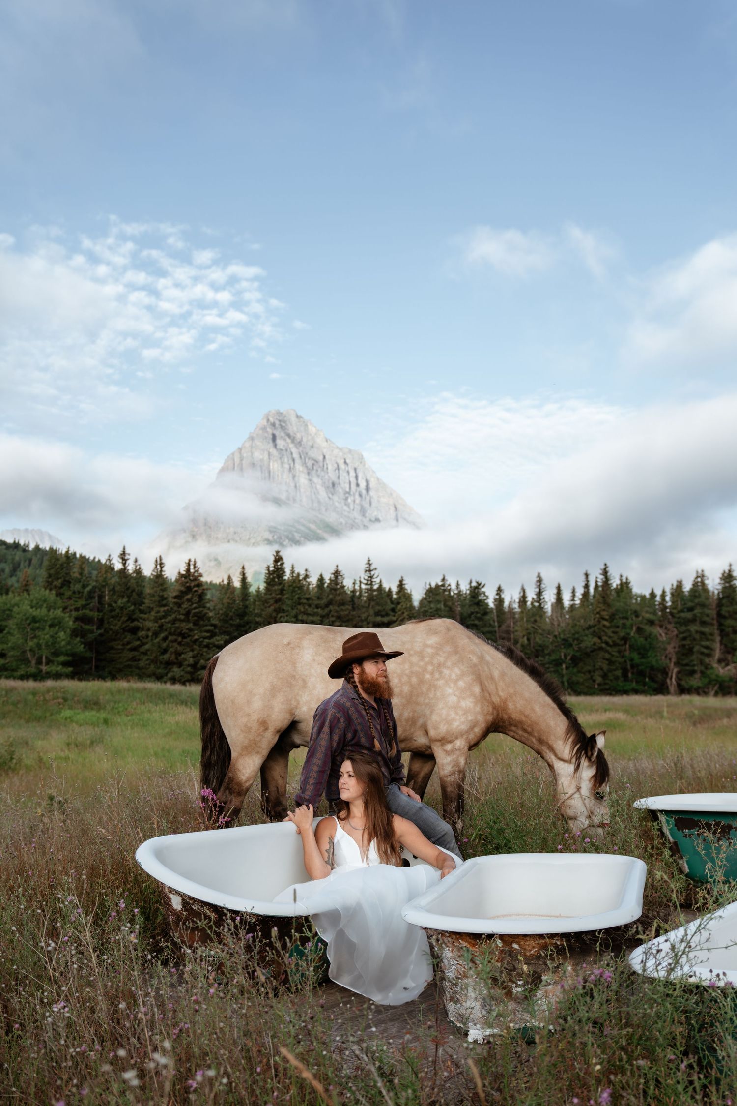Western bride and groom sit in abandoned bathtubs in large grassy field with buckskin horse and mountain behind them.