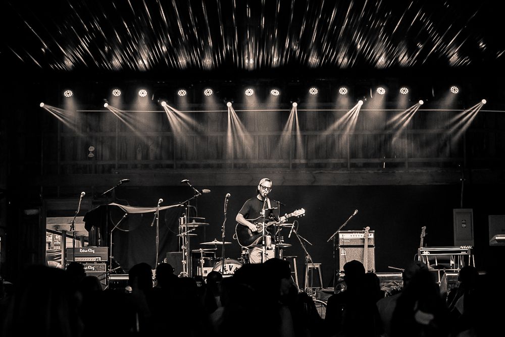 A black and white concert photo shows musicians performing under dramatic stage lighting.