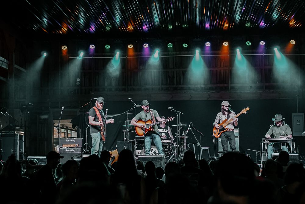 A band performs under colorful stage lights with guitars and drums on a concert stage.