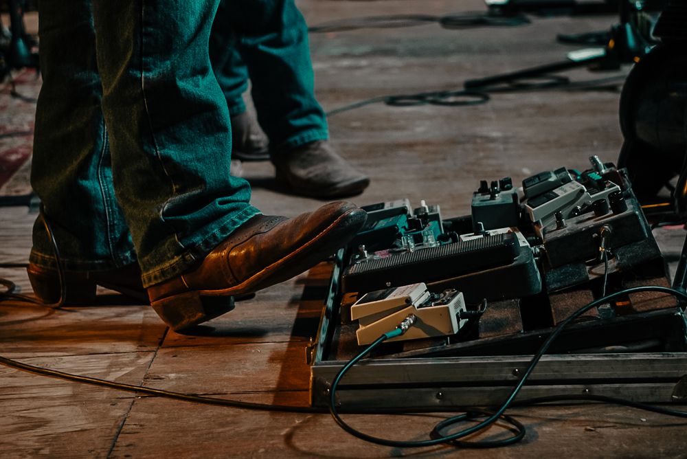 A close-up shot of guitar pedals and boots on a wooden stage floor.
