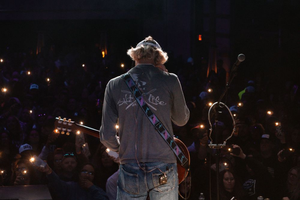 Musician performing on stage silhouetted against crowd holding lit phones.