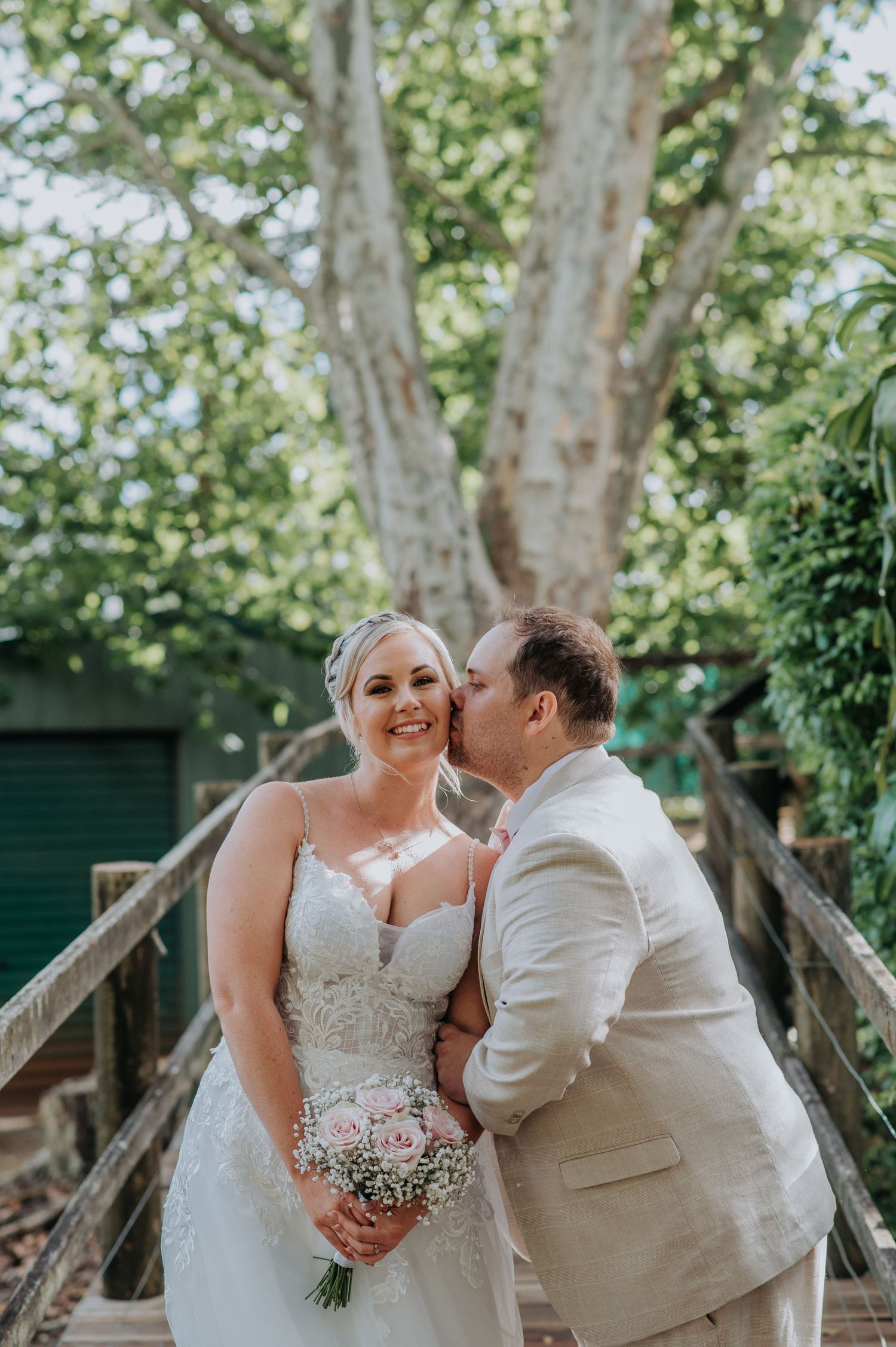 A tender wedding moment captured on a wooden bridge beneath towering trees.