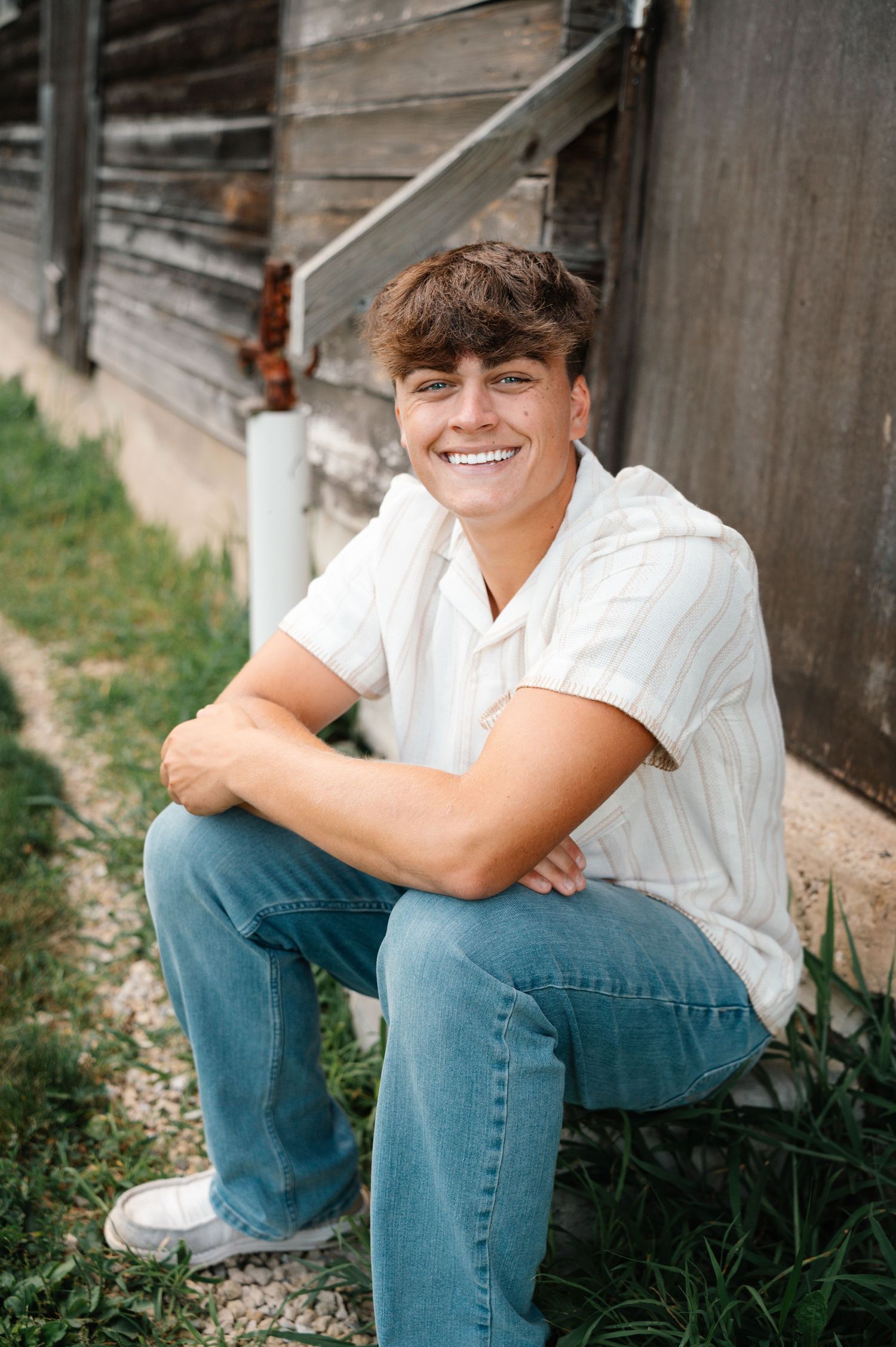 Smiling teenage boy sitting outdoors in jeans and white shirt for senior portrait photos.