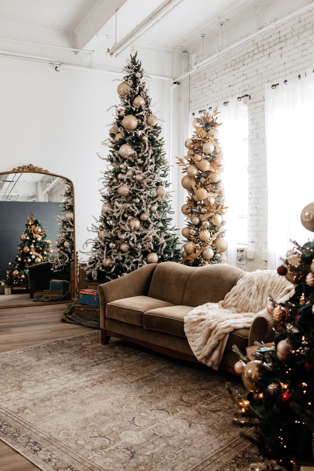 Multiple decorated Christmas trees line a bright white studio space with a brown couch and cozy throw blanket.