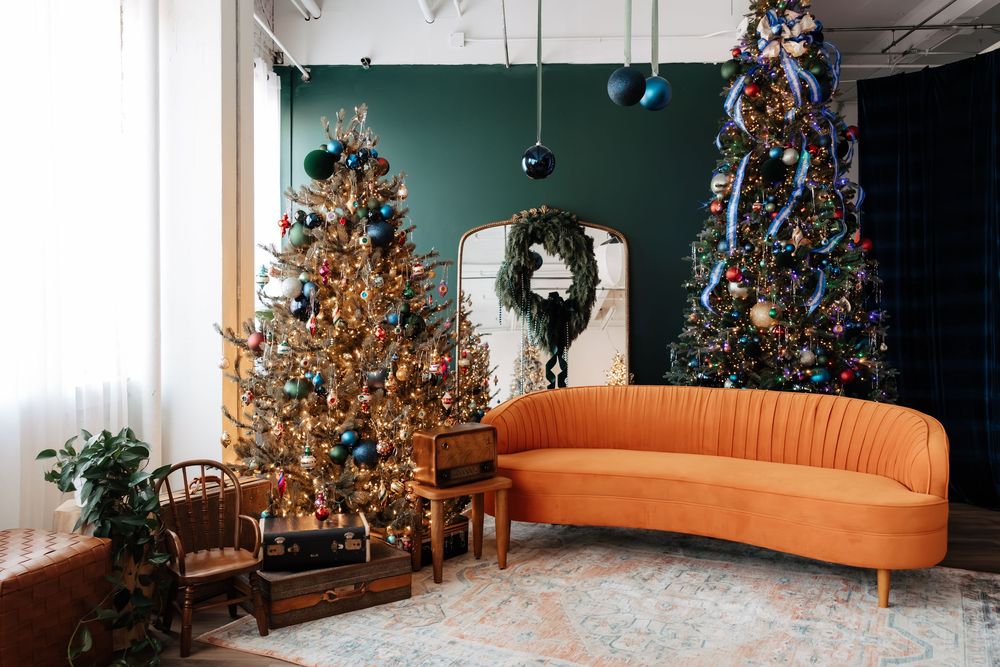 Cozy living room with two decorated Christmas trees, a curved orange velvet sofa, a green wall and a festive wreath on a mirror.