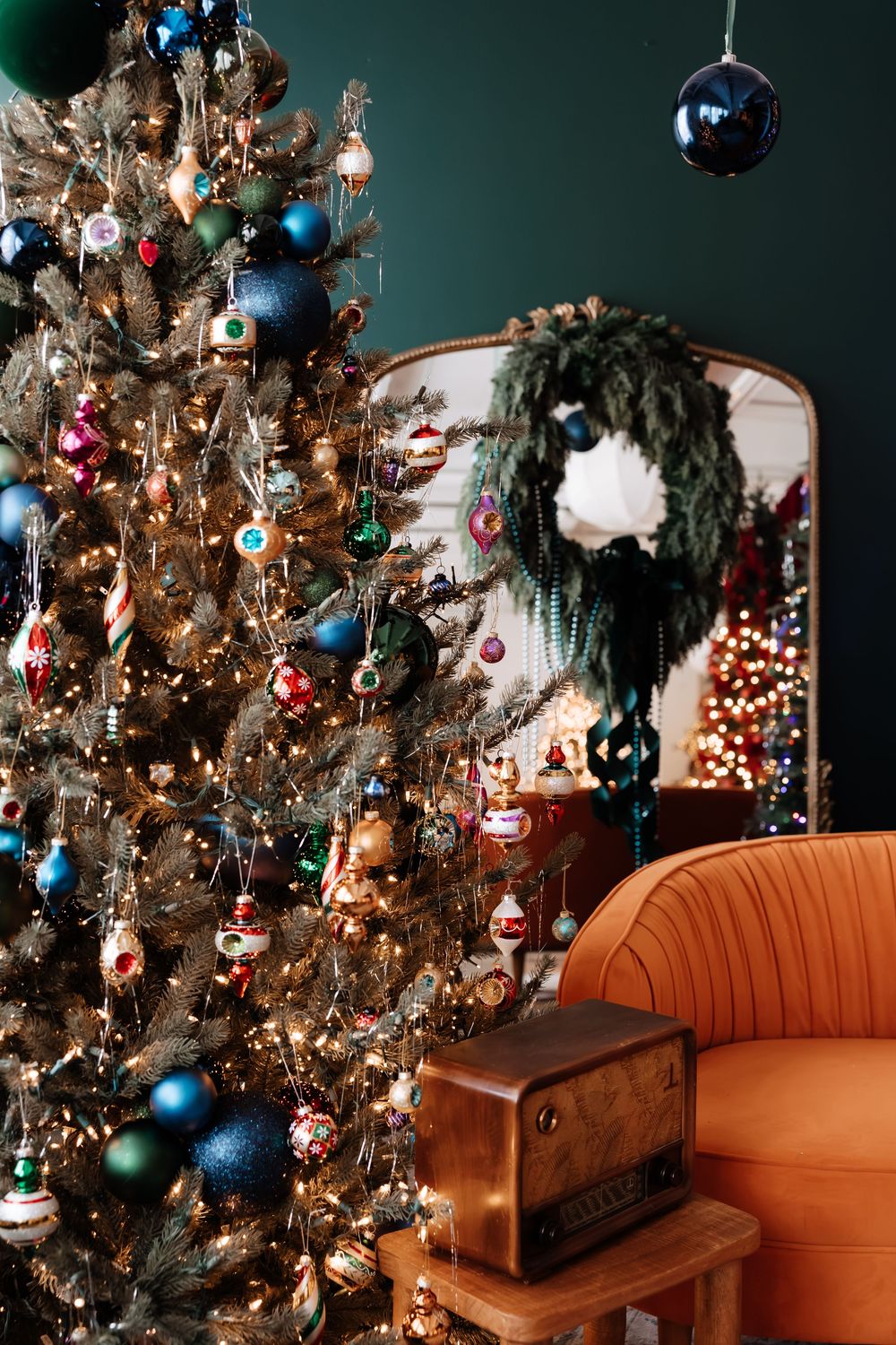 Vintage-inspired Christmas tree with colorful glass ornaments beside a velvet orange armchair, retro radio and a mirror reflecting a wreath.