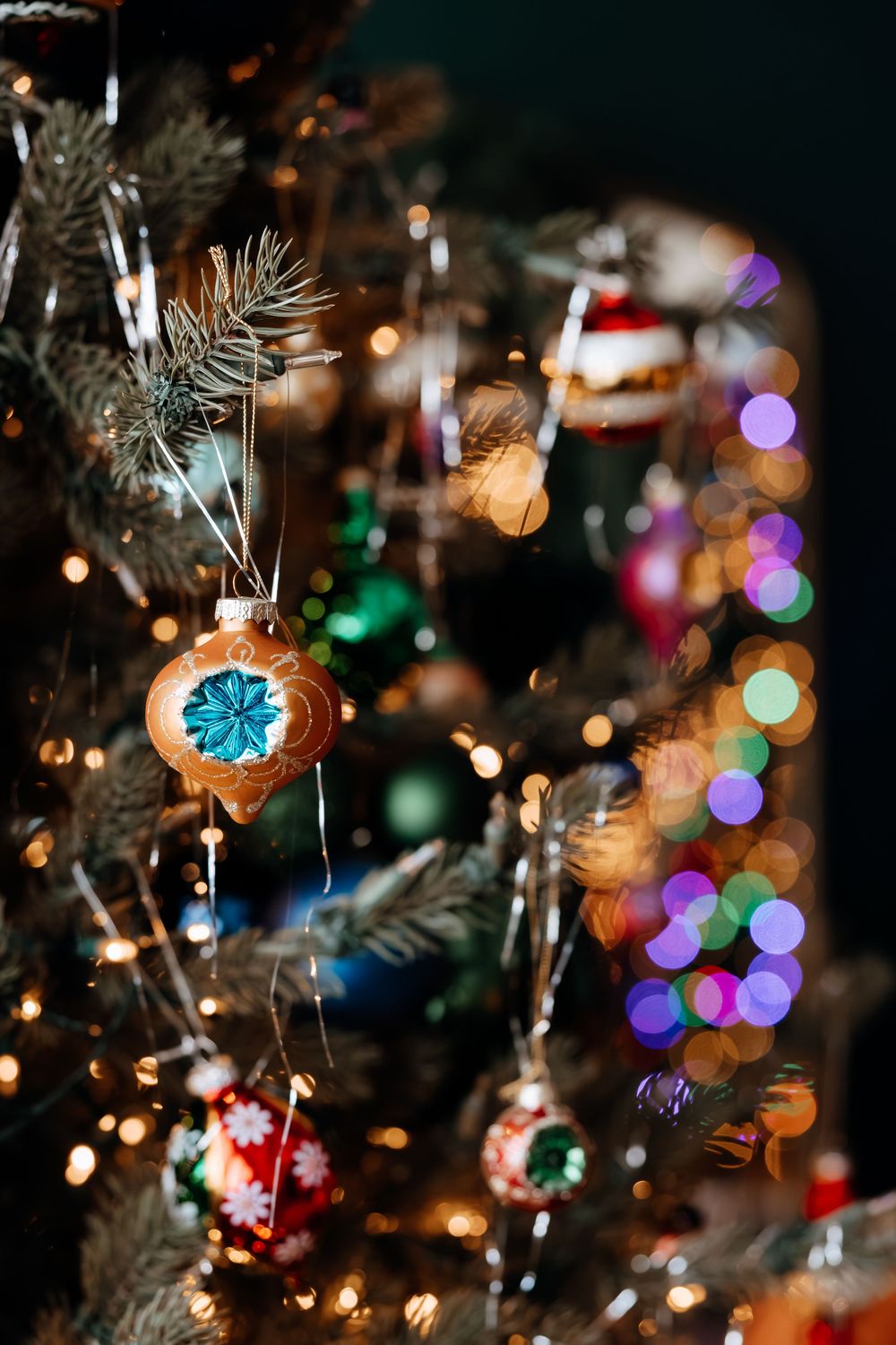 Vintage glass ornaments with teal snowflake designs hang on a Christmas tree with tinsel and softly blurred multicolored holiday lights.