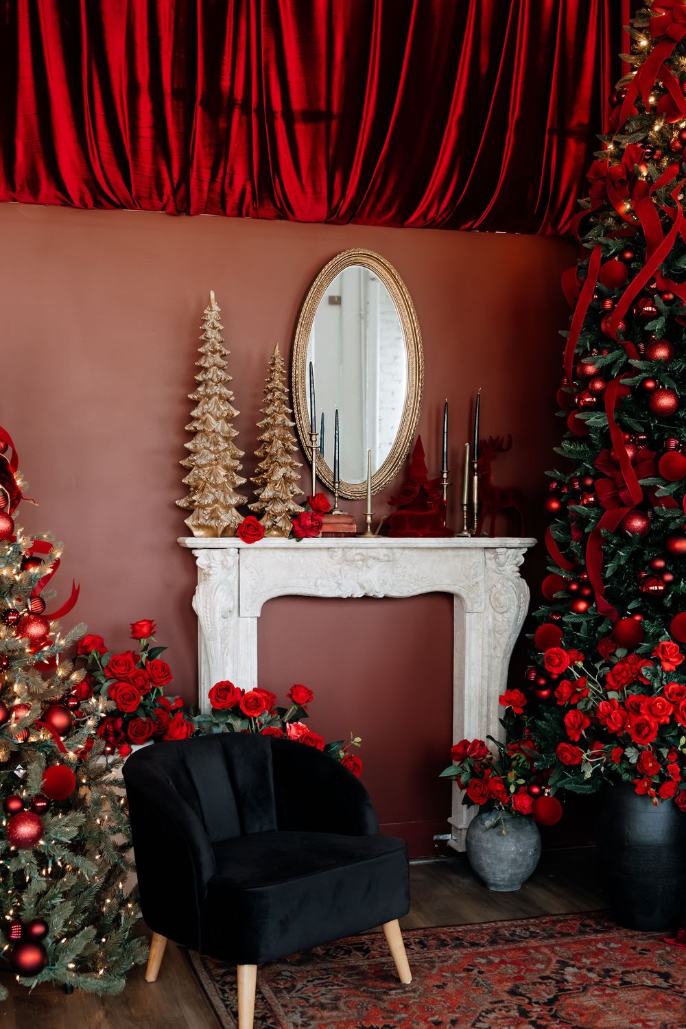 Elegant Christmas room with red velvet curtain, decorated trees, white fireplace, oval mirror, black armchair and red roses.