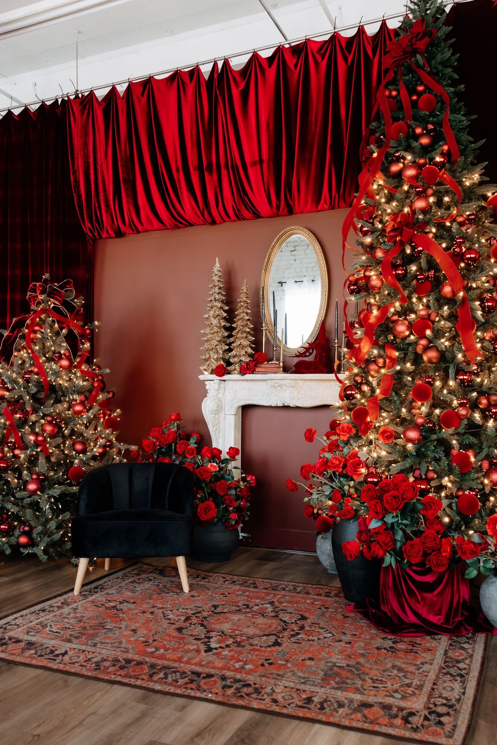 Red-themed Christmas studio with decorated trees, red velvet drapes, a black armchair, white mantel, oval mirror and vintage rug.