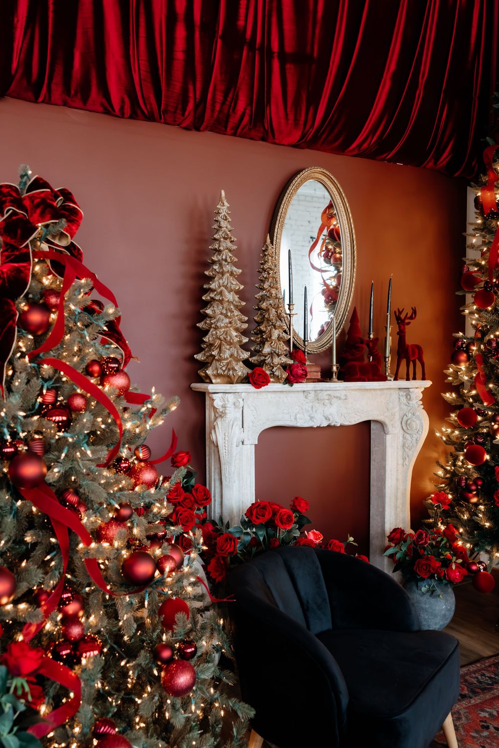 Cozy Christmas living room with decorated trees, red ornaments, white marble fireplace, gold mirror, and a black velvet armchair.