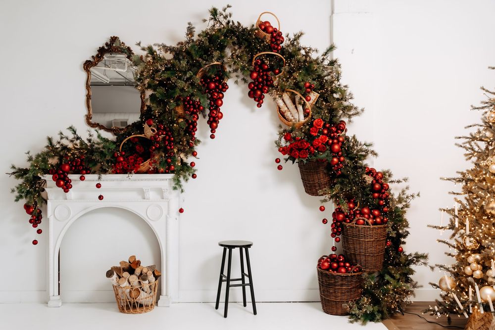 White mantel with a Christmas garland arch of evergreens and red berries, wicker baskets of ornaments, a black stool, and a gold tree.