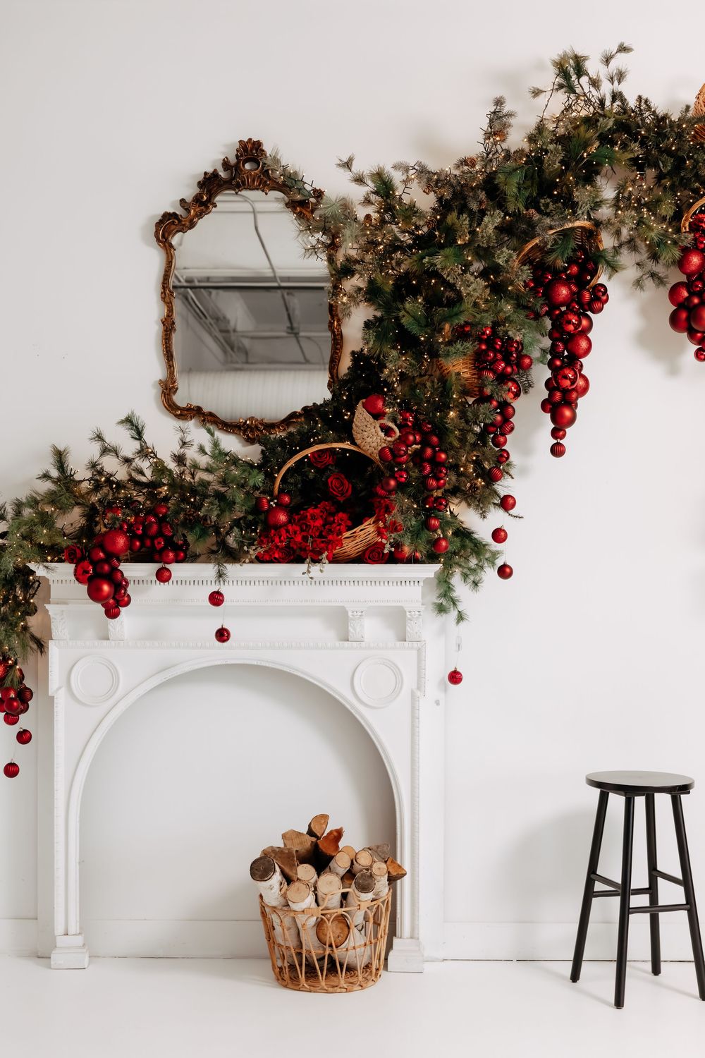 White fireplace mantel with a lush Christmas garland of red ornaments and lights, an ornate gold mirror, birch log basket and black stool.