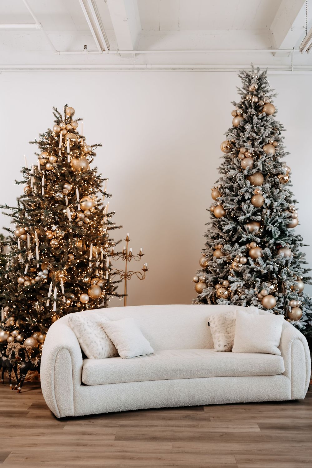 Two decorated Christmas trees with gold ornaments and candles flank a cream bouclé sofa with pillows in a bright, modern studio.