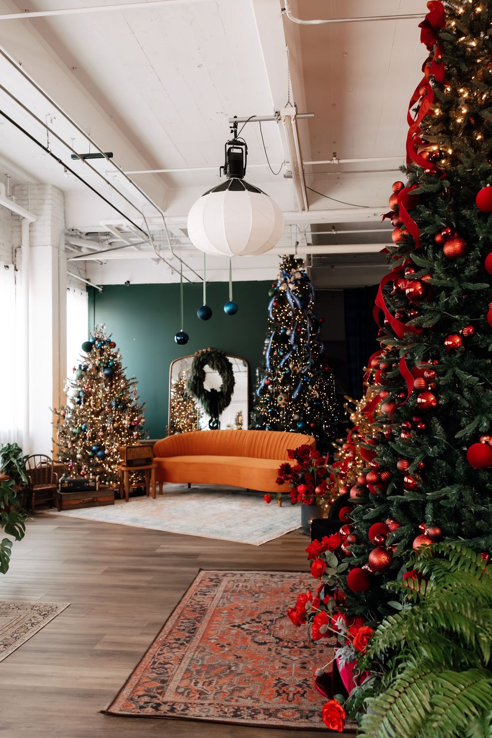 Festive studio with a red-ribboned Christmas tree, orange velvet sofa, patterned rug, wreath, and lit trees against a green wall.