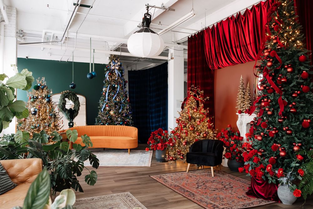 Festively decorated loft studio with red-ornamented Christmas trees, red velvet curtains, an orange sofa, plants and a vintage rug.