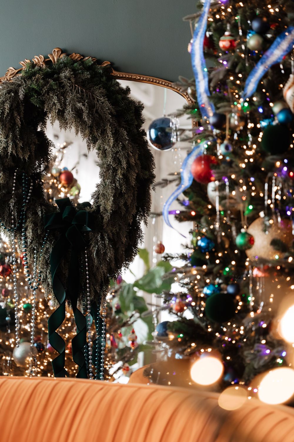 Decorated Christmas tree with blue, red and green ornaments and warm lights beside a lush evergreen wreath with ribbon and beads.