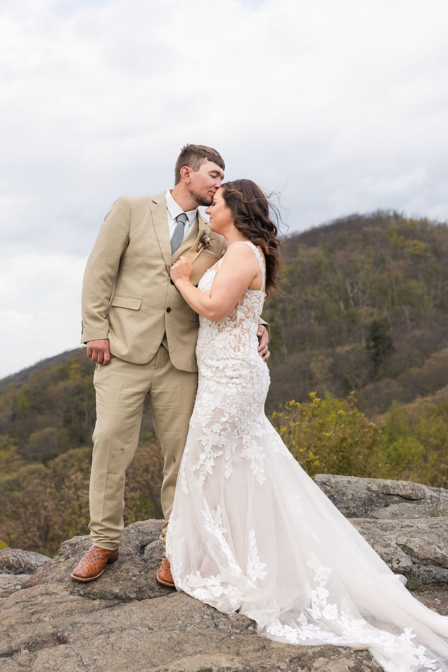 Bride and groom share a romantic kiss on a rocky overlook with mountain views on their wedding day.