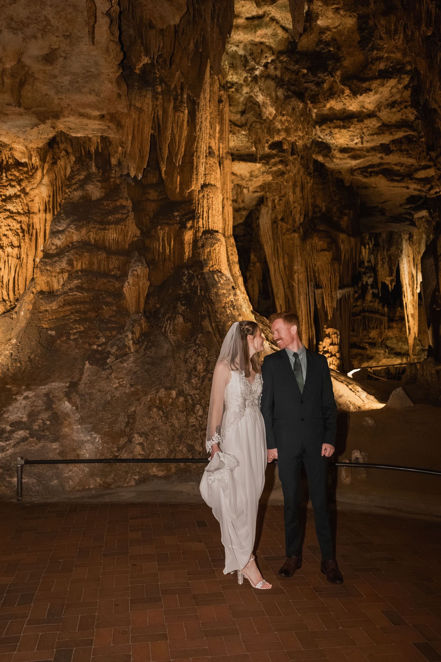 A couple stands together in a dimly lit cave with dramatic stalactites and rock formations illuminated from below.