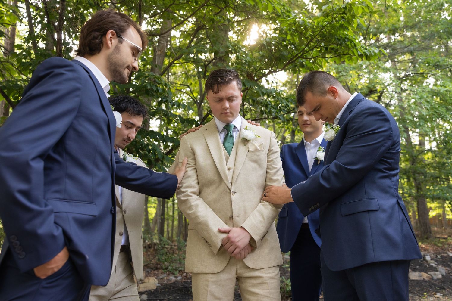 Groomsmen in navy suits pray with groom in tan suit outdoors before wedding ceremony in wooded setting.