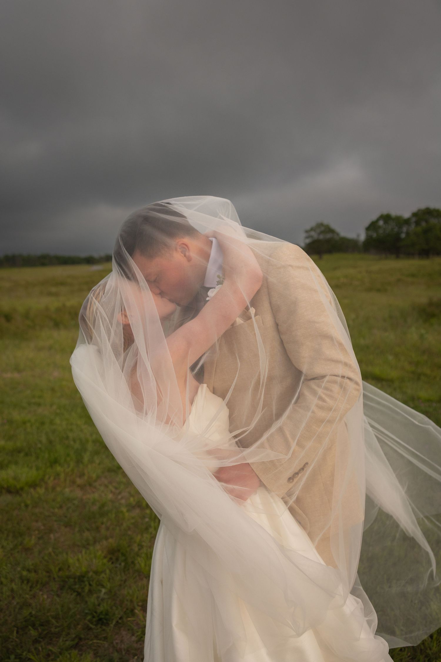 Bride and groom sharing a romantic kiss under a flowing veil in a green field with dramatic stormy skies.