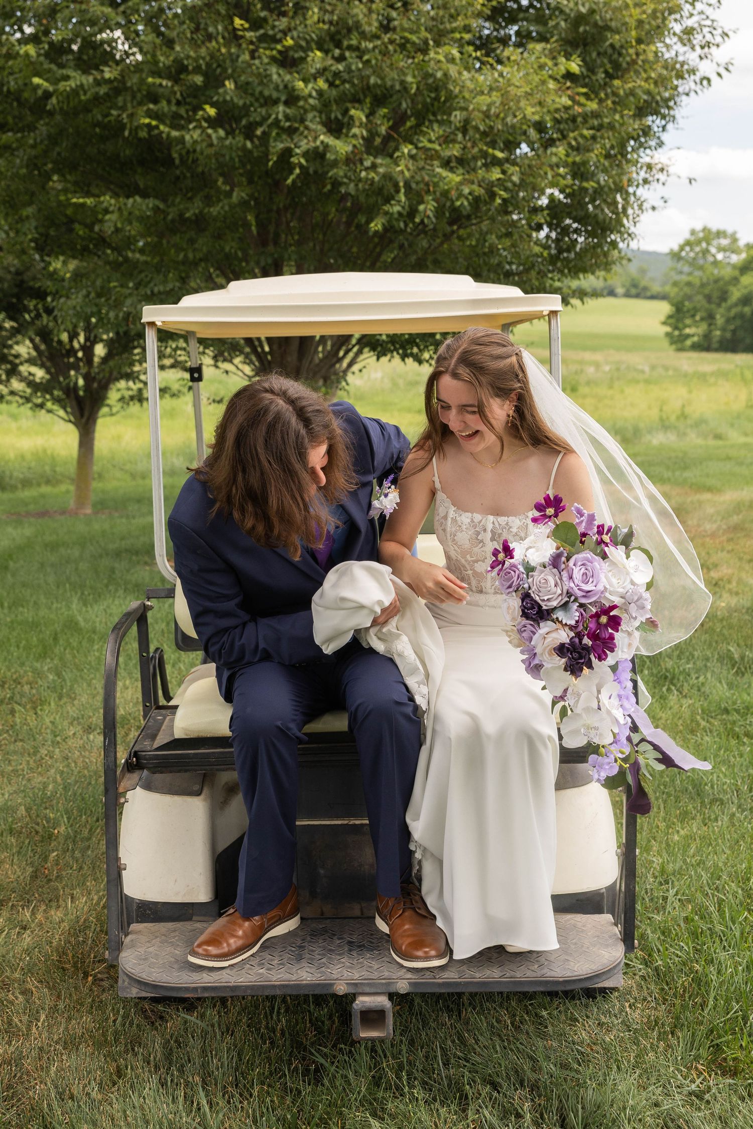 Bride and groom sitting on back of golf cart, bride holding purple bouquet, smiling together outdoors.