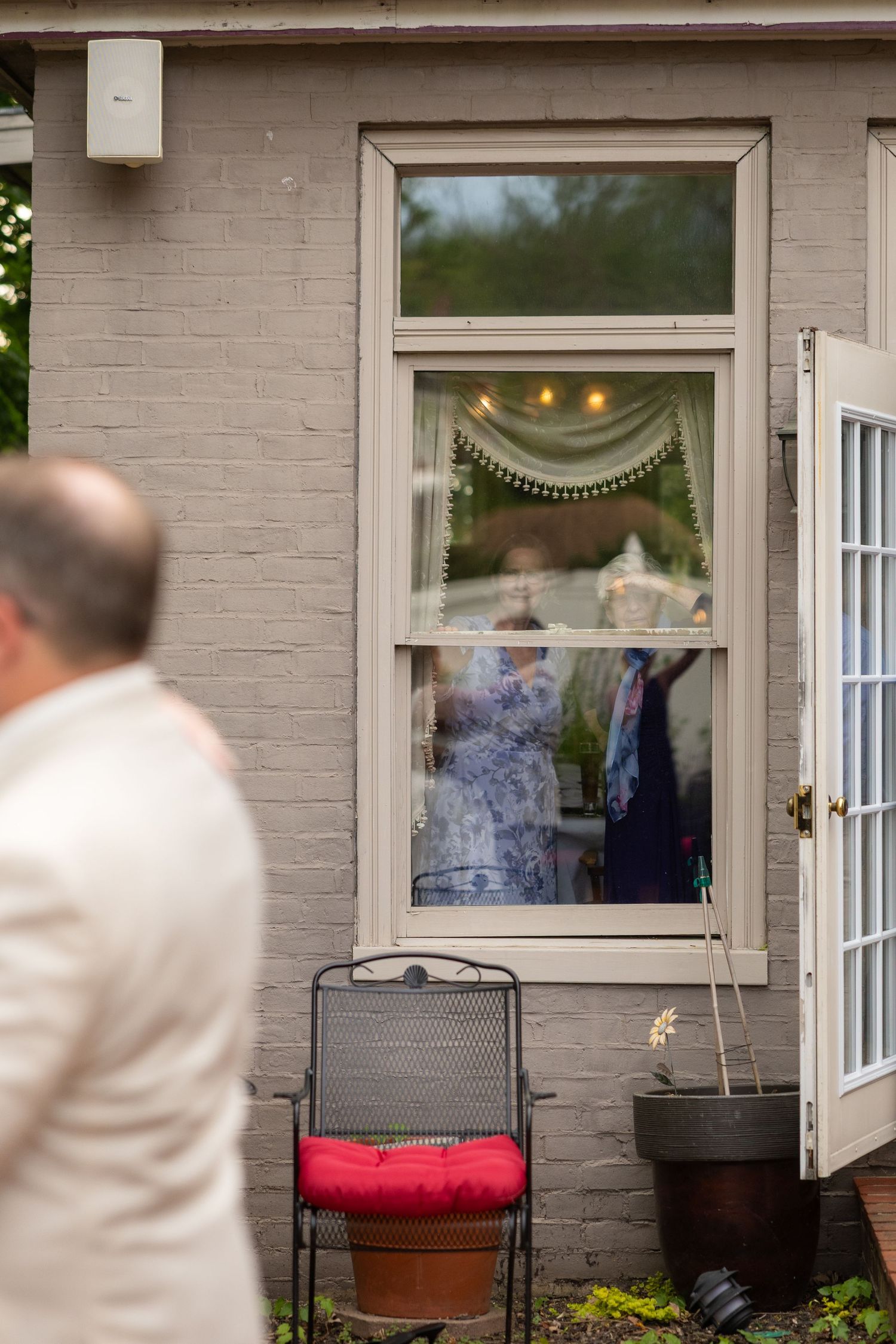 People visible through a window of a gray brick building with a chair and potted plant outside.