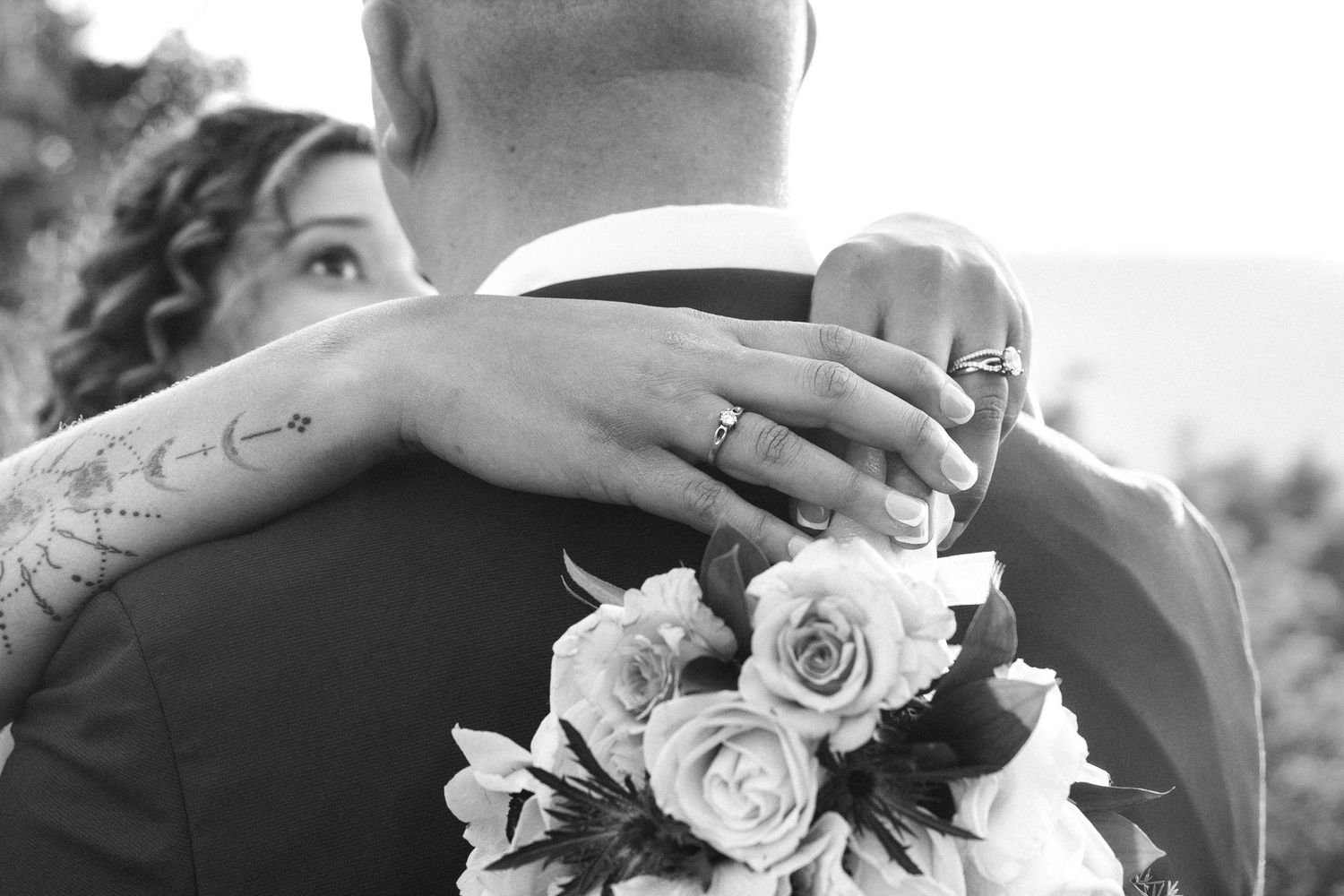 Black and white wedding photo of bride holding roses bouquet, arm around groom, showcasing wedding ring and floral tattoo.