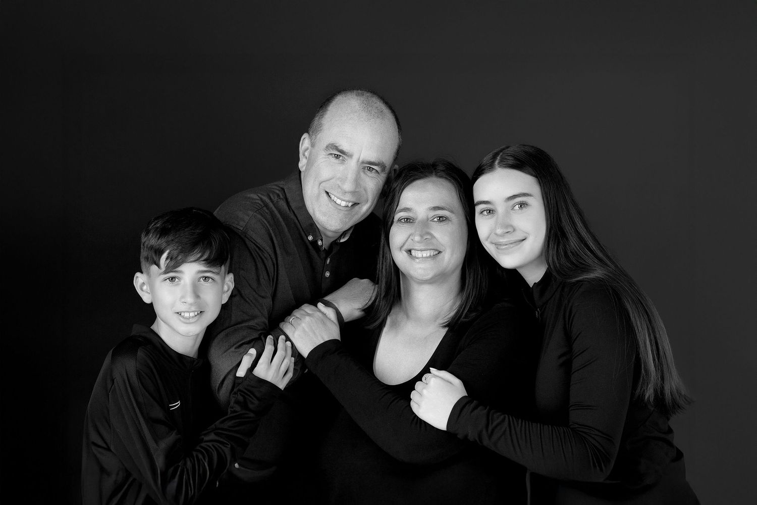 A family with two young children sits together for a casual portrait against a gray studio backdrop.